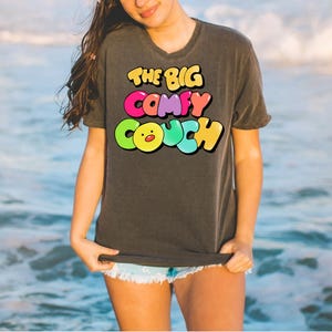 K&ouml;nnte beinhalten: Ein dunkelgraues T-Shirt mit dem Text "THE BIG COMFY COUCH" in bunten Blasenbuchstaben. Die Buchstaben sind schwarz umrandet und das "O" in "COUCH" ist ein gelbes Cartoon-K&uuml;ken mit rotem Schnabel.
