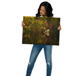 May include: A woman holds a framed photograph of a lake with a small cabin on the water. The lake is surrounded by trees with fall foliage in shades of red, orange, and yellow. The water reflects the colors of the trees.