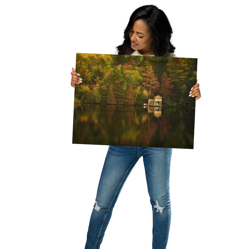 May include: A woman holds a framed photograph of a lake with a small cabin on the water. The lake is surrounded by trees with fall foliage in shades of red, orange, and yellow. The water reflects the colors of the trees.
