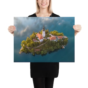 Puede incluir: Una impresi&oacute;n en p&oacute;ster que presenta una vista a&eacute;rea de una peque&ntilde;a isla con una iglesia y edificios rodeados por un lago. La iglesia tiene un alto campanario blanco y un tejado rojo. La isla est&aacute; cubierta de &aacute;rboles verdes y el agua es azul oscuro.