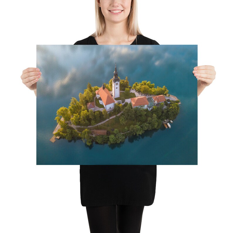Puede incluir: Una impresi&oacute;n en p&oacute;ster que presenta una vista a&eacute;rea de una peque&ntilde;a isla con una iglesia y edificios rodeados por un lago. La iglesia tiene un alto campanario blanco y un tejado rojo. La isla est&aacute; cubierta de &aacute;rboles verdes y el agua es azul oscuro.