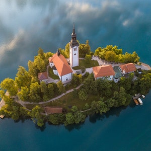Puede incluir: Vista a&eacute;rea de una peque&ntilde;a isla con una iglesia y edificios, rodeada por un lago. La iglesia tiene una alta torre de campana blanca y un tejado rojo. La isla est&aacute; cubierta de &aacute;rboles y vegetaci&oacute;n verdes. El agua es de color azul profundo.