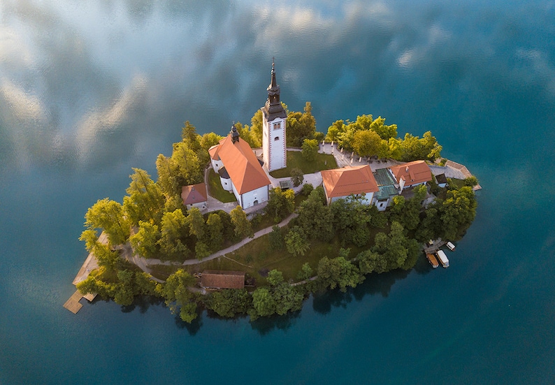 Puede incluir: Vista a&eacute;rea de una peque&ntilde;a isla con una iglesia y edificios, rodeada por un lago. La iglesia tiene una alta torre de campana blanca y un tejado rojo. La isla est&aacute; cubierta de &aacute;rboles y vegetaci&oacute;n verdes. El agua es de color azul profundo.