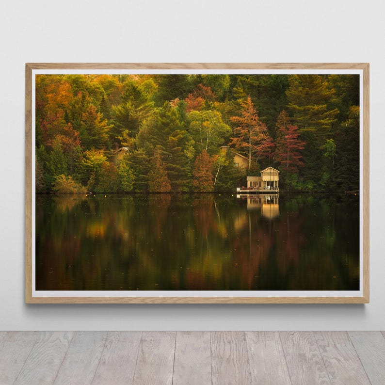May include: A photograph of a lake with a small wooden cabin on the shore. The lake is surrounded by trees with fall foliage in shades of red, orange, yellow, and green. The water is calm and reflects the colors of the trees.