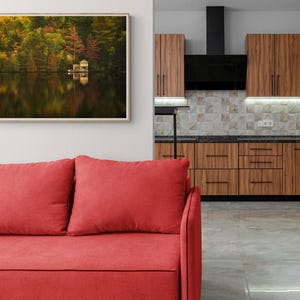 May include: A framed photograph of a lake with trees in the background and a small cabin on the water. The lake is reflecting the trees and the sky. The photo is hanging on a white wall above a red sofa.
