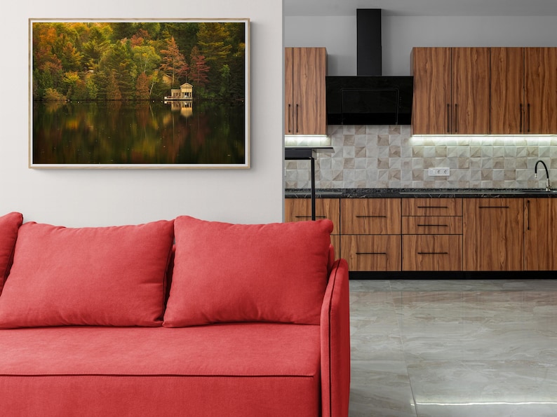 May include: A framed photograph of a lake with trees in the background and a small cabin on the water. The lake is reflecting the trees and the sky. The photo is hanging on a white wall above a red sofa.