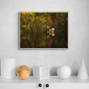 May include: A photograph of a small wooden cabin on a lake surrounded by trees with fall foliage. The cabin is reflected in the still water.