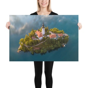 Puede incluir: Una impresi&oacute;n en p&oacute;ster que presenta una vista a&eacute;rea pintoresca de una peque&ntilde;a isla con una iglesia y edificios rodeados por un lago. La iglesia tiene un alto campanario y un tejado rojo. La isla est&aacute; cubierta de &aacute;rboles verdes y el agua es azul oscuro.