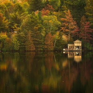 May include: A wooden cabin with a porch sits on the edge of a lake surrounded by trees with fall foliage. The lake is calm and reflects the trees and cabin.
