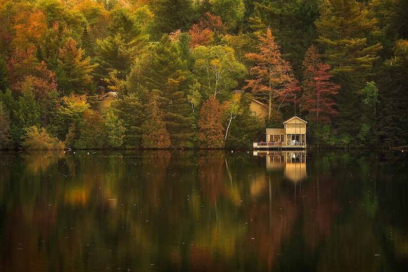 May include: A wooden cabin with a porch sits on the edge of a lake surrounded by trees with fall foliage. The lake is calm and reflects the trees and cabin.