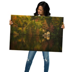 May include: A photograph of a lake with a small cabin on the water. The lake is surrounded by trees with fall foliage in shades of red, orange, yellow, and green. The water reflects the colors of the trees.