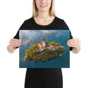 Puede incluir: Un p&oacute;ster con una vista a&eacute;rea de una isla con una iglesia y edificios rodeados de agua. La iglesia tiene un alto campanario blanco y un tejado rojo. La isla est&aacute; cubierta de &aacute;rboles verdes y rodeada de agua azul.