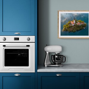 Puede incluir: Fotograf&iacute;a enmarcada de un paisaje de una peque&ntilde;a isla con una iglesia y edificios, rodeada de agua. La fotograf&iacute;a se muestra en una pared azul claro en un entorno de cocina. Se ven un horno blanco y armarios azules.