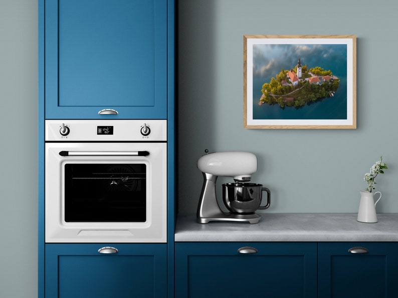 Puede incluir: Fotograf&iacute;a enmarcada de un paisaje de una peque&ntilde;a isla con una iglesia y edificios, rodeada de agua. La fotograf&iacute;a se muestra en una pared azul claro en un entorno de cocina. Se ven un horno blanco y armarios azules.