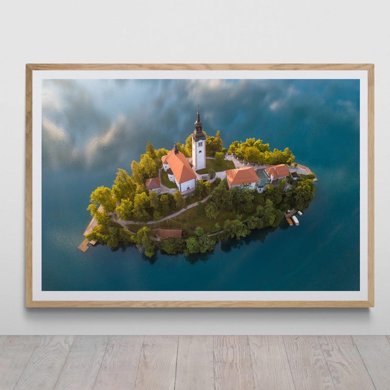 Puede incluir: Vista a&eacute;rea de una peque&ntilde;a isla con una iglesia y varios edificios rodeados por un lago. La iglesia tiene una alta torre de campana blanca y un tejado rojo. La imagen est&aacute; enmarcada en un marco de madera clara.