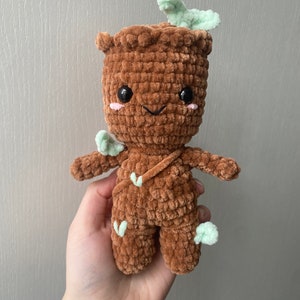 Puede incluir: Un juguete de peluche de color marrón tejido a crochet con una cara sonriente y hojas verdes. El juguete es un personaje de la película Guardianes de la Galaxia.
