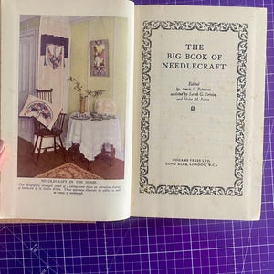 May include: An open book titled "The Big Book of Needlecraft" with illustrations of needlecraft in a home setting. The book's pages display text and images of needlework, showcasing a vintage aesthetic.