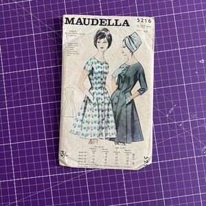 May include: A vintage sewing pattern titled "MAUDELLA" for a dress, featuring illustrations of two women in dresses. The pattern includes bust and hip measurements, and the number 5216 is visible. The background is a purple grid.
