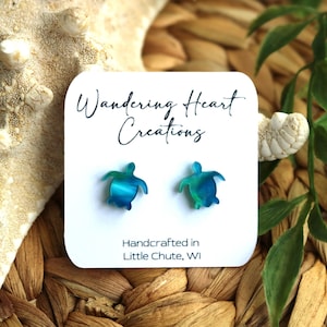 Może przedstawiać: Para kolczyków w kształcie żółwia w kolorze turkusowym i zielonym na białej kartce z tekstem "Wandering Heart Creations" i "Handcrafted in Little Chute, WI".