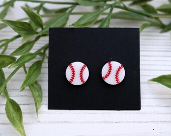 Peut inclure: Une paire de boucles d'oreilles à clous sur le thème du baseball. Chaque boucle d'oreille est un cercle blanc avec des coutures rouges, ressemblant à une balle de baseball. Les boucles d'oreilles sont présentées sur une carte carrée noire.