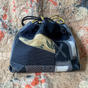 Può includere: Una borsa con coulisse realizzata con vari pannelli di tessuto nei toni del nero, grigio e oro. L'esterno presenta un mix di texture, inclusi motivi geometrici e un motivo a bambù. È chiusa da cordoncini intrecciati neri e dorati.