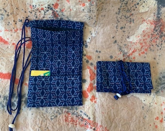 May include: A dark blue fabric pouch with a repeating geometric pattern and a long drawstring cord, featuring a small yellow card tucked into a front pocket, alongside a matching folded rectangular fabric case with a blue cord wrap.