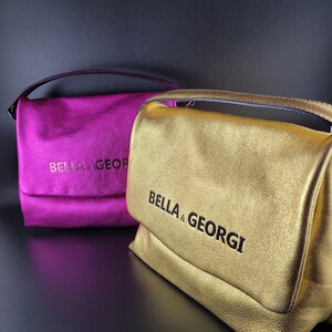 Könnte beinhalten: Zwei kleine Handtaschen, eine goldfarben und eine magentafarben, mit dem Markennamen "BELLA & GEORGI" auf der Vorderseite geprägt.