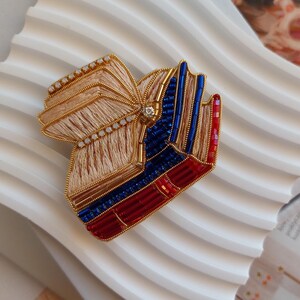 Pode incluir: Um broche artesanal em forma de pilha de três livros. O livro superior tem páginas de fio bege, enquanto os livros do meio e de baixo apresentam contas de vidro azuis e vermelhas. Um fio metálico dourado contorna as bordas do design.
