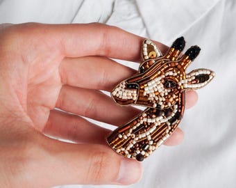 Peut inclure: Une broche en forme de girafe en perles de couleur marron, blanche et dorée. La tête de la girafe est tournée sur le côté et ses yeux sont faits de perles noires.
