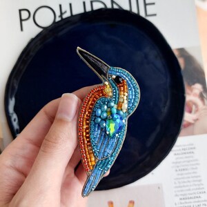 Pu&ograve; includere: Un broche a forma di uccello realizzato con perline, con uno schema di colori blu, arancio e verde. L'uccello &egrave; fatto di piccole perline e ha un becco lungo e sottile. Il broche &egrave; attaccato a uno spillo di metallo.
