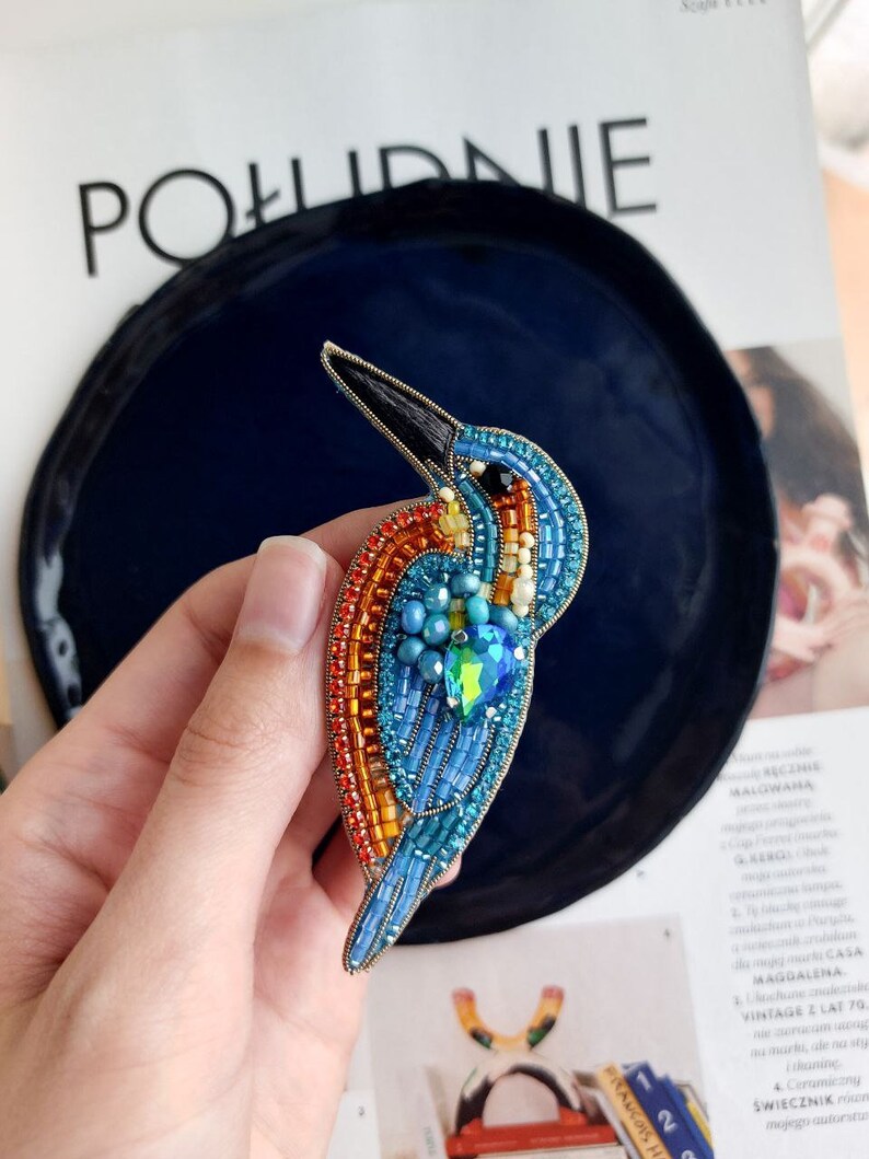 Pu&ograve; includere: Un broche a forma di uccello realizzato con perline, con uno schema di colori blu, arancio e verde. L'uccello &egrave; fatto di piccole perline e ha un becco lungo e sottile. Il broche &egrave; attaccato a uno spillo di metallo.