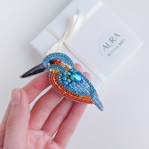 Pu&ograve; includere: Un broche a forma di uccello in perline con accenti blu, arancio e verde. Il broche &egrave; fatto di perline e ha un design dettagliato. Il broche &egrave; su uno sfondo bianco.