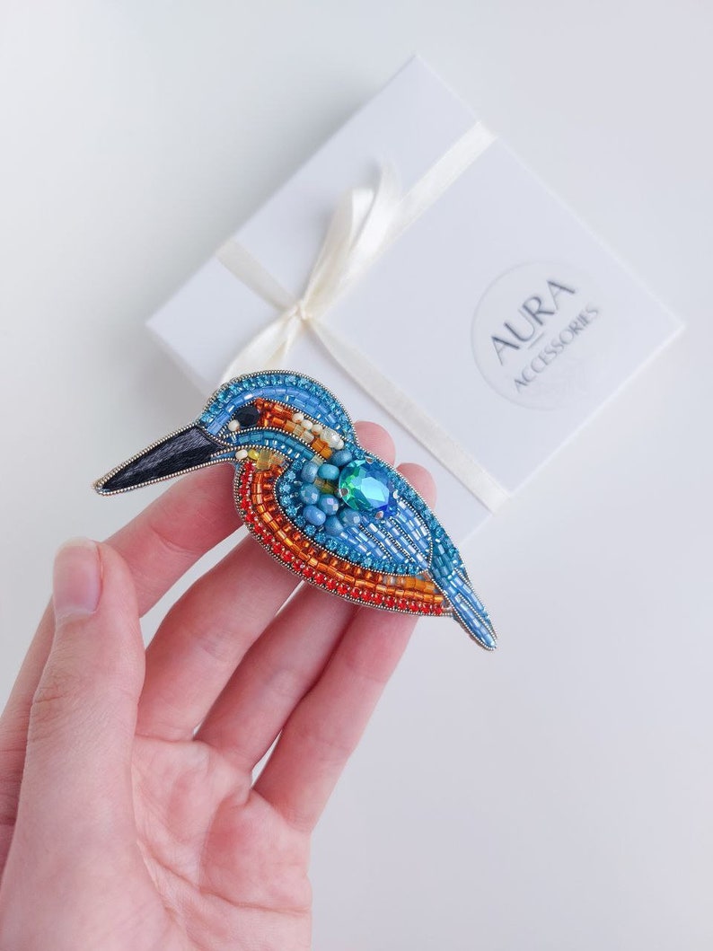 Pu&ograve; includere: Un broche a forma di uccello in perline con accenti blu, arancio e verde. Il broche &egrave; fatto di perline e ha un design dettagliato. Il broche &egrave; su uno sfondo bianco.