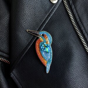 Pu&ograve; includere: Una spilla a forma di uccello realizzata con perline, in tonalit&agrave; di blu, arancio e giallo, con un solo occhio di pietra preziosa verde, appuntata su una giacca di pelle nera.