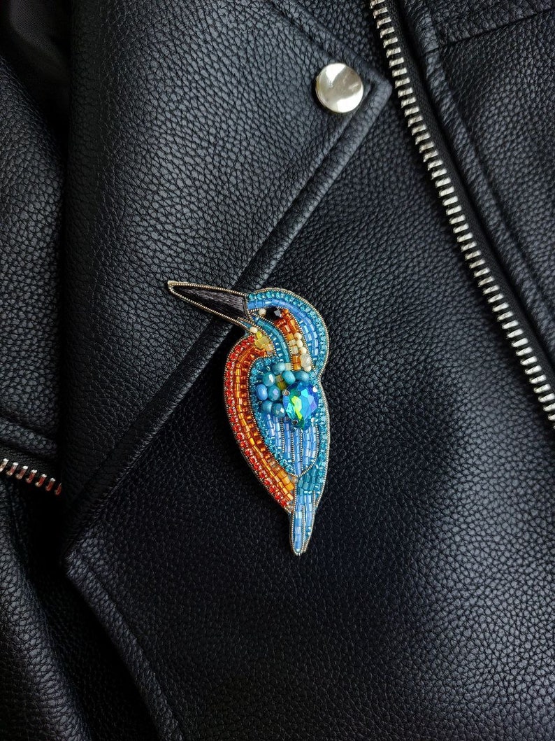 Pu&ograve; includere: Una spilla a forma di uccello realizzata con perline, in tonalit&agrave; di blu, arancio e giallo, con un solo occhio di pietra preziosa verde, appuntata su una giacca di pelle nera.