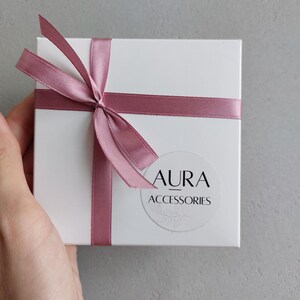 Pu&ograve; includere: Una scatola regalo bianca con un nastro rosa legato a fiocco. La scatola ha un adesivo rotondo con il testo "AURA ACCESSORIES" su di essa.