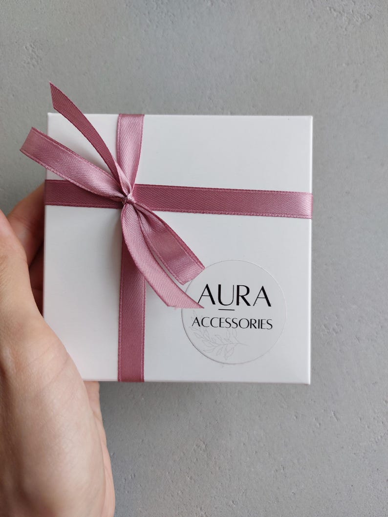 Pu&ograve; includere: Una scatola regalo bianca con un nastro rosa legato a fiocco. La scatola ha un adesivo rotondo con il testo "AURA ACCESSORIES" su di essa.