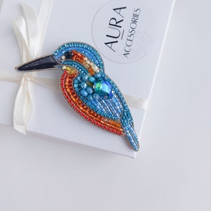 Pu&ograve; includere: Una spilla a forma di uccello realizzata con perline, con accenti blu, arancioni e verdi. La spilla ha la forma di un martin pescatore ed &egrave; appoggiata su una scatola bianca con la scritta "AURA ACCESSORIES" sopra.