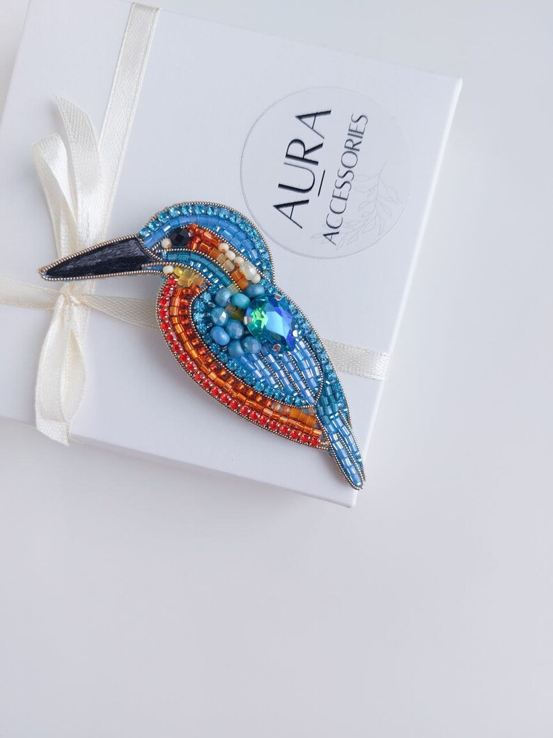 Pu&ograve; includere: Una spilla a forma di uccello realizzata con perline, con accenti blu, arancioni e verdi. La spilla ha la forma di un martin pescatore ed &egrave; appoggiata su una scatola bianca con la scritta "AURA ACCESSORIES" sopra.