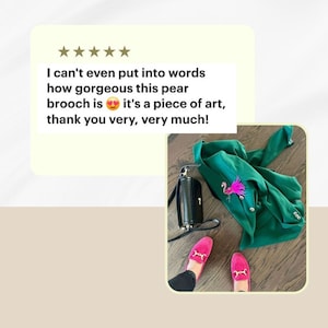 Pu&ograve; includere: Un broche a forma di fenicottero rosa con dettagli scintillanti, indossato su una giacca verde. La giacca &egrave; appoggiata su un pavimento di legno con una borsa nera e mocassini rosa.