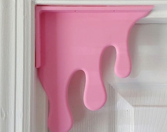 May include: Pink plastic shelf bracket shaped like a drip. The bracket has a sharp corner and a smooth, glossy finish.
