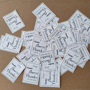 Peut inclure: Plusieurs petites cartes rectangulaires blanches éparpillées sur une surface marron. Chaque carte affiche le mot "Merci" dans une police stylisée, avec des traductions en plusieurs langues, dont "Thanks" et "Gracias."