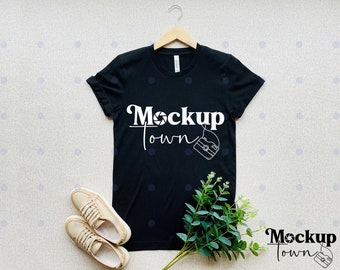 Puede incluir: Camiseta negra con el texto "Mockup Town" y un gráfico de cámara. La camiseta está colgada de una percha de madera y hay dos zapatillas de lona beige y una planta verde en la imagen.