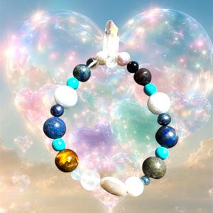 May include: A circular beaded bracelet featuring a variety of stones in shades of blue, white, brown, and metallic gray, accented with small turquoise-colored spacers and a single clear, pointed crystal charm at the top of the strand.