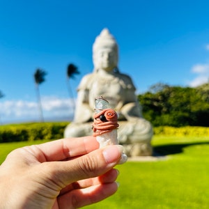 May include: A white crystal wrapped in brown leather with black beads and a silver wire. The crystal is being held in a hand with a blurred image of a white Buddha statue in the background.