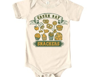 May include: Cream-colored baby bodysuit with a green and yellow graphic. The design features the words "GREEN BAY SNACKERS" above and below a collection of illustrated snacks like pretzels, cupcakes, and cheese. The bodysuit has short sleeves and a round neckline.