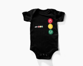 May include: Black baby onesie with a white graphic of a traffic light with three lights. The top light is red, the middle light is yellow, and the bottom light is green. The text "stink-1-2" is printed below the traffic light.