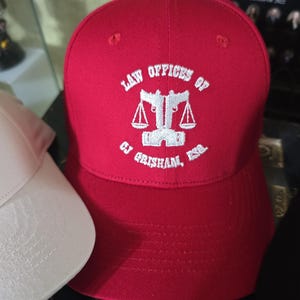 May include: A red baseball cap with the text "LAW OFFICES OF CJ GRISHAM, ESQ." in white. The cap features a white emblem of scales. A white cap is partially visible on the left.