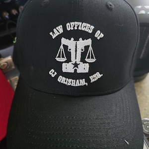 May include: Black baseball cap with white embroidered text and a logo. The text reads "LAW OFFICES OF CJ GRISHAM, ESQ." The logo features scales of justice and two crossed pistols. The cap is made of a durable fabric.