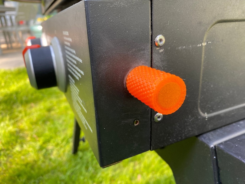 Puede incluir: Primer plano de una parrilla de gas negra con una perilla naranja. La perilla tiene una superficie texturizada y est&aacute; hecha de pl&aacute;stico.